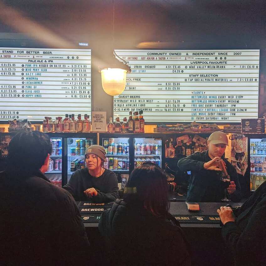 A busy bar with staff serving drinks and large beer menus overhead.