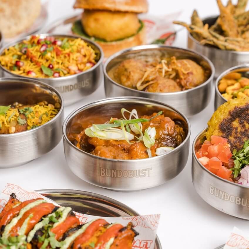 A colourful spread of Indian street food served in stainless steel bowls, including curries, chaat, and fried snacks.