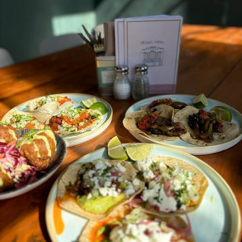 A wooden table with sunlit plates of tacos, lime wedges, and a drinks menu in the background.