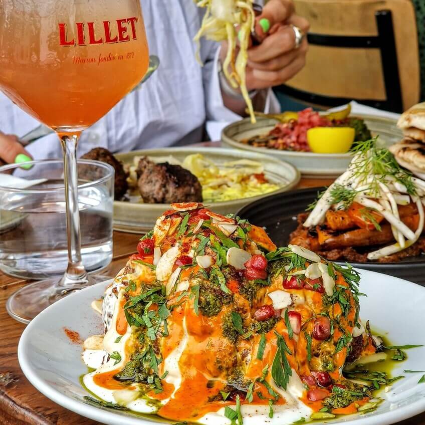 A vibrant dish of roasted cauliflower with sauce, nuts, and herbs, accompanied by cocktails and other Middle Eastern-inspired plates.