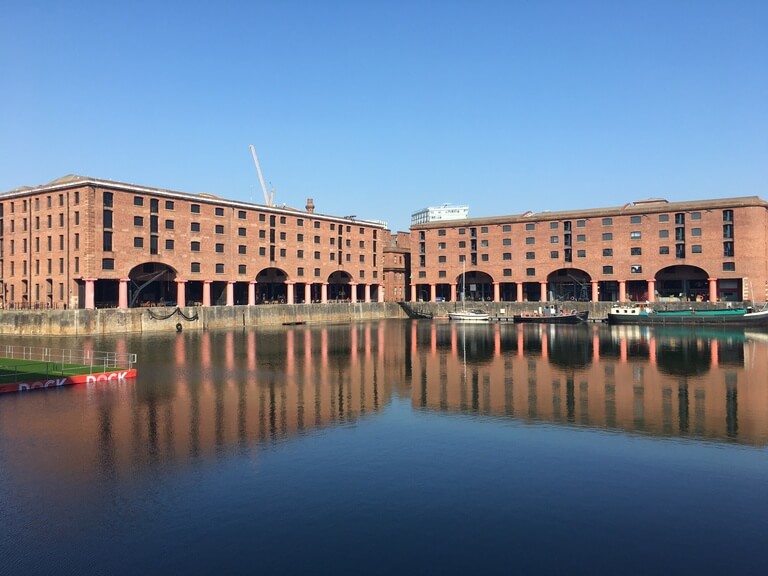 a large historic red-brick warehouse complex reflected in the calm waters of the dock, with arched openings and moored boats