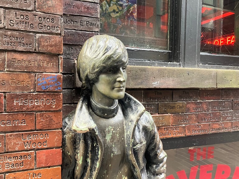 a bronze statue of John Lenon leaning against a brick wall with band names inscribed, located on Mathew Street in Liverpool.